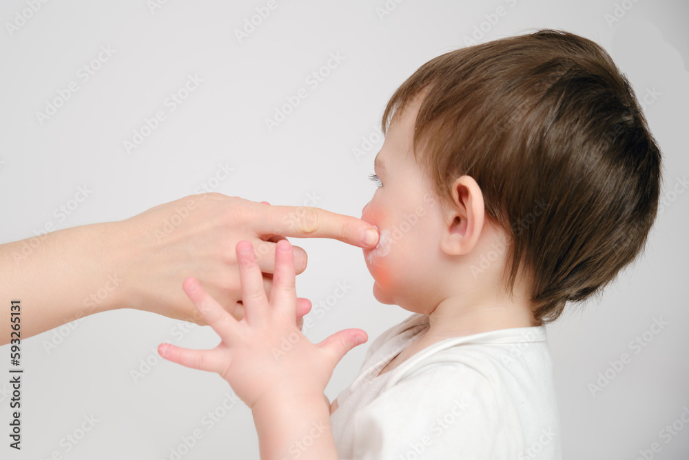 Woman mother smears her cheeks with baby cream on a studio white background. Mom smears child's face with a moisturizer for eczema. Kid about two years old (one year nine months) Woman mother smears her cheeks with baby cream on a studio white background. Mom smears child's face with a moisturizer for eczema. Kid about two years old (one year nine months)