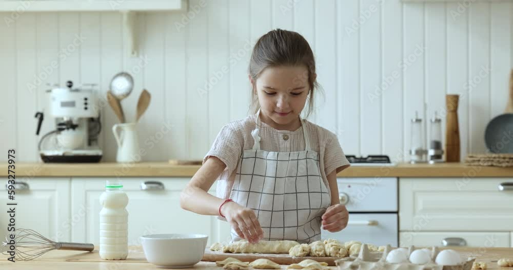 Cute little kid girl wearing apron, baking in home kitchen, kneading, shaping dough on floury table with bakery food ingredients, enjoying dessert meal preparation, culinary hobby Cute little kid girl wearing apron, baking in home kitchen, kneading, shaping dough on floury table with bakery food ingredients, enjoying dessert meal preparation, culinary hobby