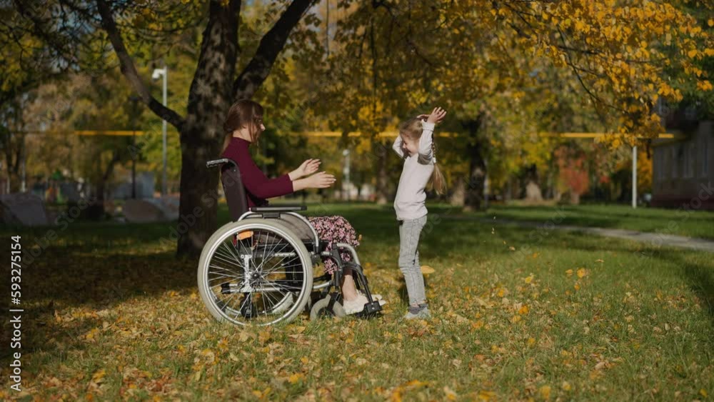 Young mother with legs injury throws bunch of dry leaves up above daughter. Preschooler girl raises hands rejoicing at game in autumn park side view Young mother with legs injury throws bunch of dry leaves up above daughter. Preschooler girl raises hands rejoicing at game in autumn park side view