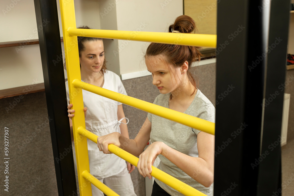 Obraz premium Disabled Teenage Girl Does Physical Exercises In Rehabilitation Room Near Gymnastic Wall. Doctor Supports Child With Special Needs. Rehabilitation. Cerebral Palsy. Motor Disorder. Horizontal plane.