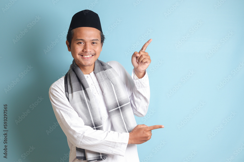 Portrait of young Asian muslim man showing product and pointing with his hand and finger to the side. Isolated image on blue background Portrait of young Asian muslim man showing product and pointing with his hand and finger to the side. Isolated image on blue background