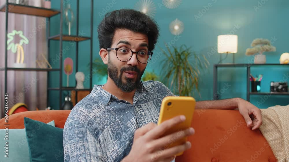 Oh my God Wow. Excited happy indian man use smartphone typing browsing celebrating success winning, lottery jackpot goal achievemen, good news, victory. Hindu guy holds mobile phone at home room