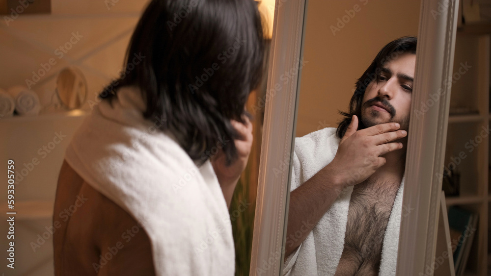 Man grooming. Beard care. Masculine beauty. Pensive handsome shirtless brunet guy with white towel touching face hair looking in mirror after shower.