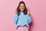 Emotional happy euphoric Brazilian woman clenches fists and celebrates successs cheers over something dressed in loose blue jumper smiles toothily isolated over pink background. Hooray I did it