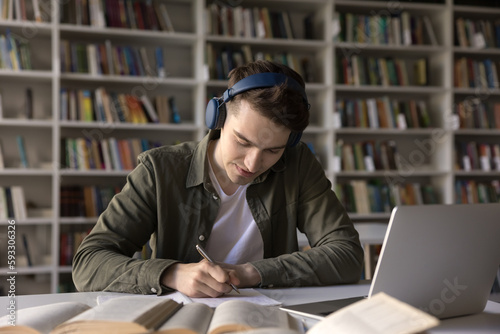 Focused student guy sit at desk work hard on research project in library, listen audio lesson through headphones improve language skills, get ready for admission, gain new knowledge use modern tech