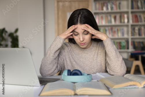 Schoolgirl sit at desk with textbook in library cramming, prepare for exams, think, solve math task, read theory, holds her head feels overworked, tired from studying or cramming. Information overload photo