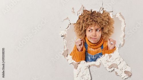 Strict displeased woman contractor with curly hair clenches fists and looks angrily aside smeared with plaster involved in repairing breaks through hole wears sweater and overalls focused aside