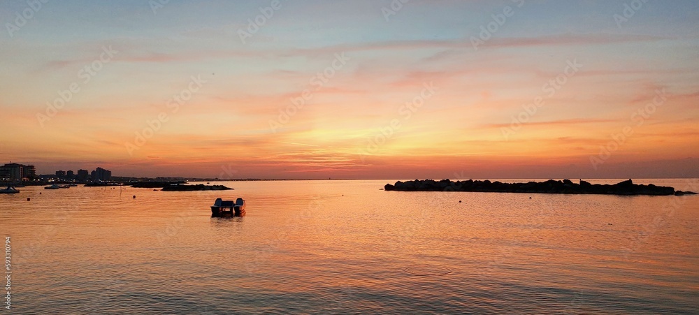Tramonto al mare, il riflesso del sole da al mare e al cielo sfumature rosse, arancio, rosa e giallo.
Il mare è calmo, tra gli scogli c'è una piccola imbarcazione, intorno calma e serenità.