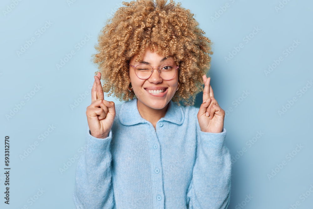 Hopeful curly haired woman crosses fingers feels superstitous believes in good luck bites lips and winks eye wears spectacles and casual jumper isolated over blue background. May my dreams come true Hopeful curly haired woman crosses fingers feels superstitous believes in good luck bites lips and winks eye wears spectacles and casual jumper isolated over blue background. May my dreams come true