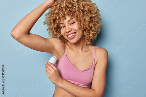 Horizontal shot of cheerful curly woman raises arm applies deodorant on armpit wears cropped top smiles pleasantly isolated over blue wall. Sporty female model puts antiperspirant stick in underarms