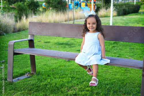 Niña pequeña chiquita bebe hermosa latina sonriente tierna amorosa jugando disfrutando y saludando a la cámara sentada en una banca divertida en un hermoso día soleado al atardecer en el parque photo
