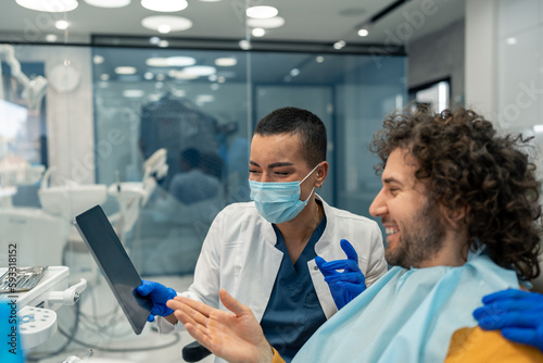 Friendly female dentist with protective face mask using digital tablet showing entertaining online presentation to a handsome satisfied male patient at modern dentist's office during appointment.