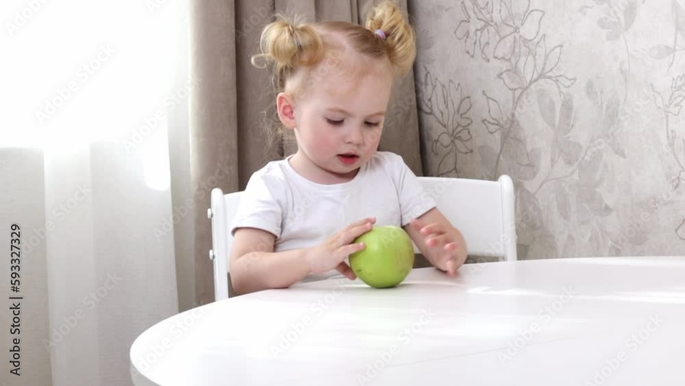 A little girl in a white t-shirt sits at a table and plays with a green apple A little girl in a white t-shirt sits at a table and plays with a green apple