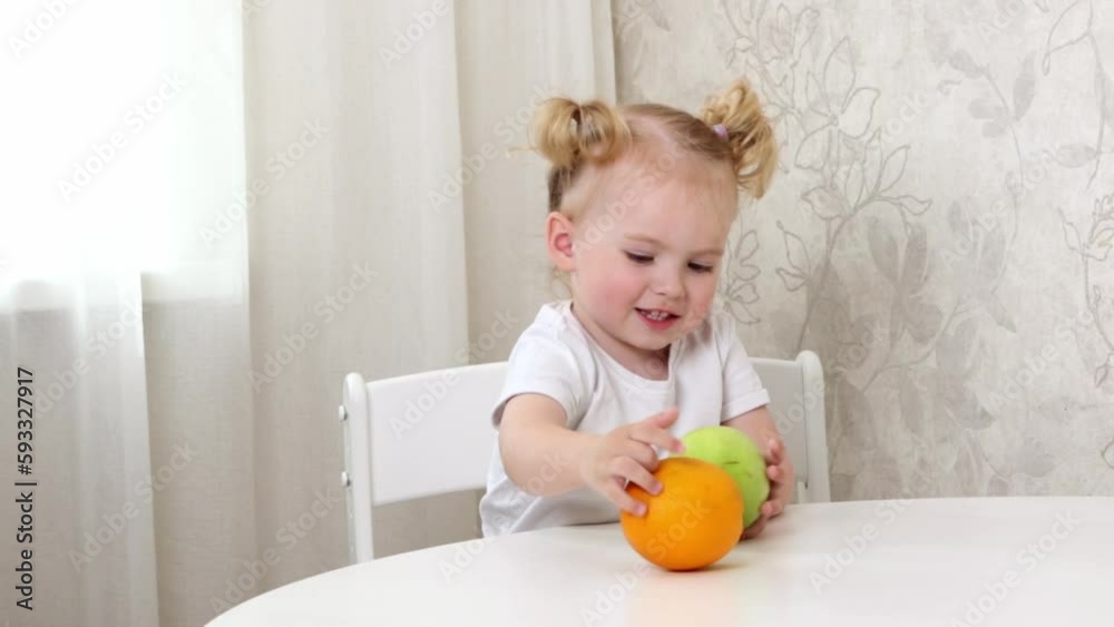 A little girl in a white T-shirt sits at the table and plays with fruits. lemon, orange and apple A little girl in a white T-shirt sits at the table and plays with fruits. lemon, orange and apple