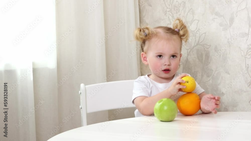 A little girl in a white T-shirt sits at the table and plays with fruits. lemon, orange and apple A little girl in a white T-shirt sits at the table and plays with fruits. lemon, orange and apple