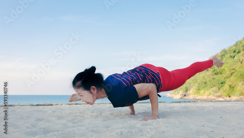 Strong Asian senior elder woman practicing yoga on the beach in summer morning with beautiful blue sky. Healthy lifestyle and fitness leisure concept.