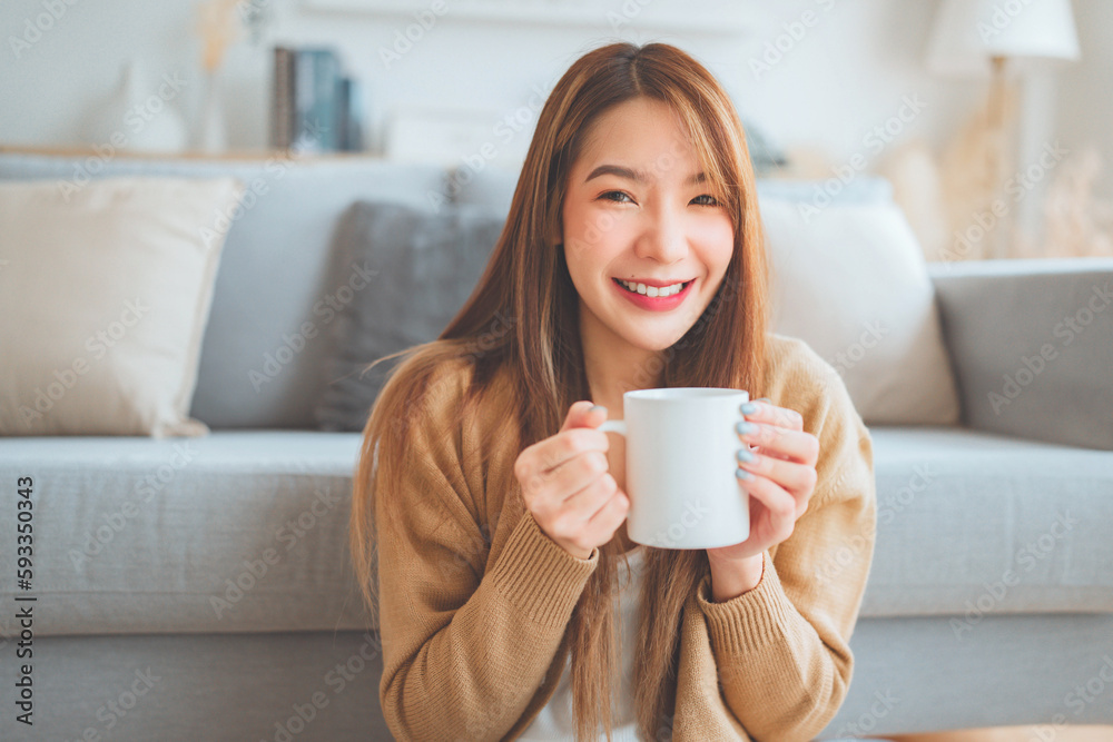 Happy young asian female enjoying a cup of coffee while sitting on the rug beside to the sofa at home, Cosy scene, Smiling pretty woman drinking hot tea in winter. Happy young asian female enjoying a cup of coffee while sitting on the rug beside to the sofa at home, Cosy scene, Smiling pretty woman drinking hot tea in winter.