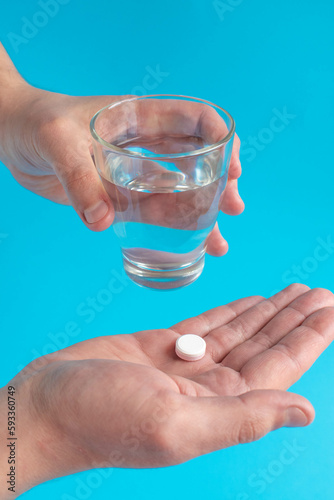 A man is holding a tablet and a glass of water over a blue background, ready to take his medicine. The sick man needs medicine for headaches and colds, a painkiller, a food supplement. 
