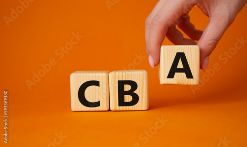 CBA - Cost Benefit Analysis symbol. Wooden cubes with word CBA. Beautiful orange background. Businessman hand. Business and Cost Benefit Analysis concept. Copy space.
