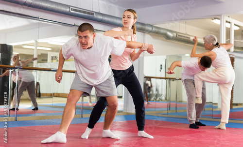 Caucasian woman performing elbow strike while sparring with man in gym during self-defence training. Senior woman training in background.