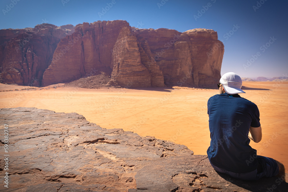 Wadi Rum w Jordanii. Mężczyzna siedzący na pustynnej skale patrzący na pustynię.  - obrazy, fototapety, plakaty 