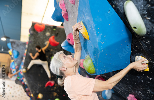 Joyful concentrated senior woman busy with her hobby bouldering, climbing on artificial wall in gym, extreme sport, rock-climbing concept