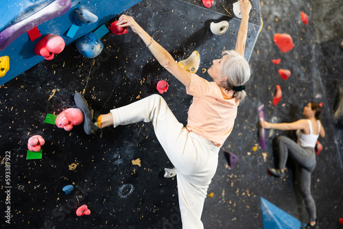 Positive focused mature woman exercising on black climbing wall without safety belts and preparing for summer mountain hiking in game center indoors