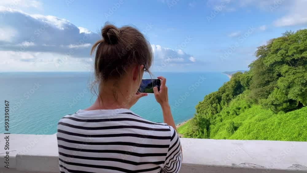 Woman in striped t shirt is holding smartphone and taking photo or shooting video of the Black Sea, blue cloudy sky, green trees in the botanical garden. Georgian landscape near Batumi in Georgia Woman in striped t shirt is holding smartphone and taking photo or shooting video of the Black Sea, blue cloudy sky, green trees in the botanical garden. Georgian landscape near Batumi in Georgia