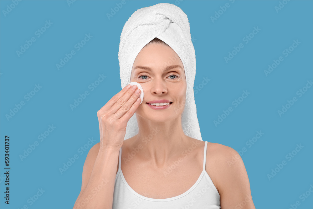 Beautiful woman in terry towel removing makeup with cotton pad on light blue background Beautiful woman in terry towel removing makeup with cotton pad on light blue background