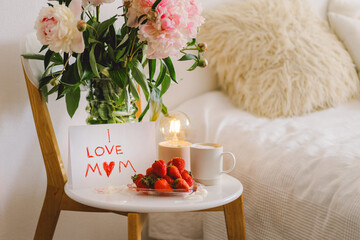 Breakfast for Mothers Day. Heart shaped white plate with fresh strawberries, cup of coffee, gift and Peonys bouquet with gift in bed. Still life composition. Happy Mother's Day.