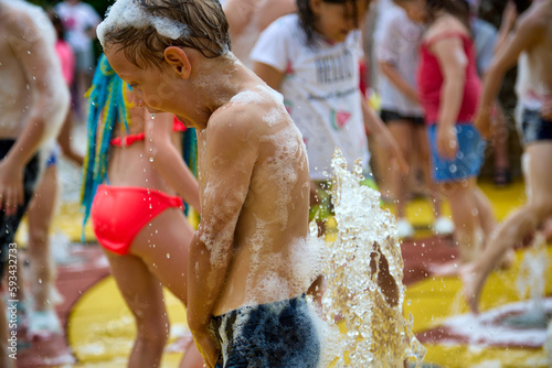Funny boy with foam on his head and body stands under the water jet of a pedestrian city fountain. A crowd of children have fun on the water playground in the amusement park