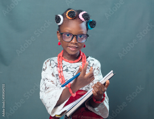 African Nigerian female girl child or student with eye glasses, wearing yoruba local dress attire and beads, holding a pen and note book while smiling and looking at the camera
