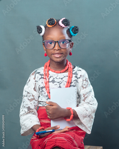 African Nigerian female girl child or student with eye glasses, wearing yoruba local dress attire and beads, holding a pen and note book while smiling and looking at the camera
