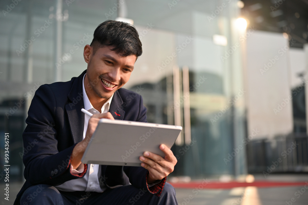 Smiling Asian businessman using his tablet while sitting on stairs outside of the company building. Smiling Asian businessman using his tablet while sitting on stairs outside of the company building.