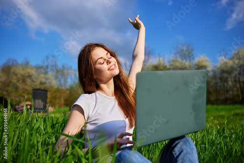 joyful woman works sitting in high grass behind a laptop raising her hand up photo