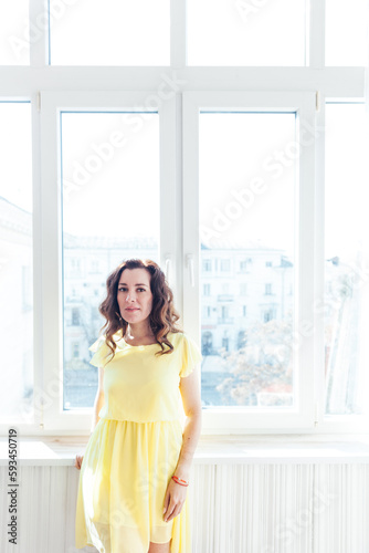 beautiful brunette woman in a yellow dress stands at a large window