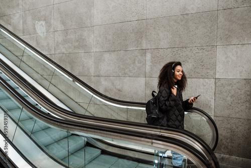 Young afro girl listening music with headphones and smartphone while going down on escalator photo