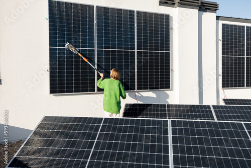 Woman washing solar panels with a brush on rooftop of her household on sunny day. Solar station care and maintenance concept