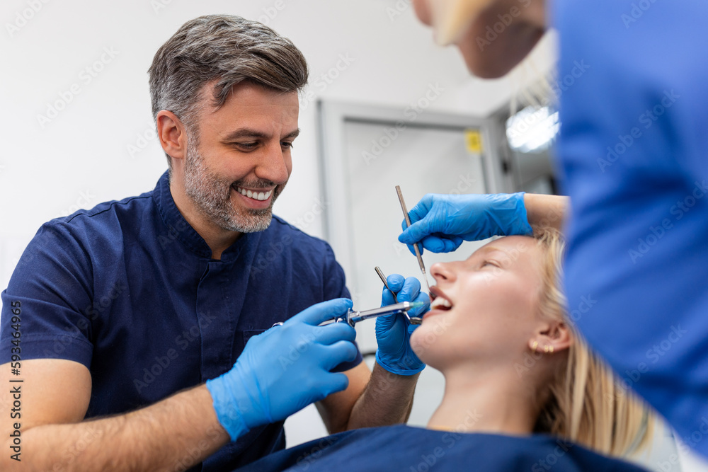 Fototapeta premium Close up of dentist's hands holding tool and syringe in hands. Pain killer in action. Female client keep mouth opened. Beautiful smile and white teeth. Cut view.