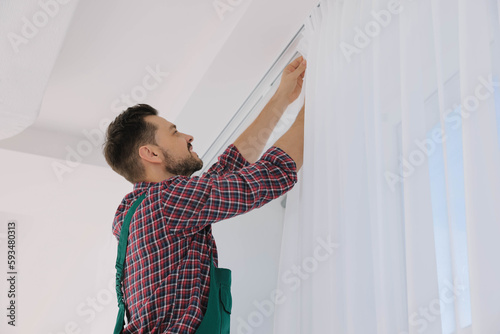 Worker in uniform hanging window curtain indoors, low angle view