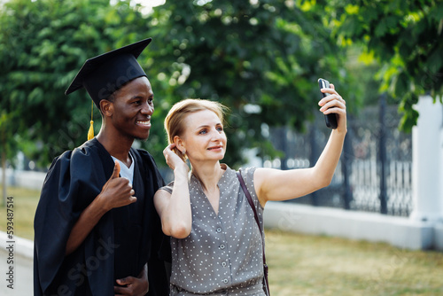 Cheerful afro american student in black graduation mantle and hat with attractive woman take selfie on smartphone. Job search, graduate from university, like, finger up, ceremonial celebration. © Юля Бурмистрова