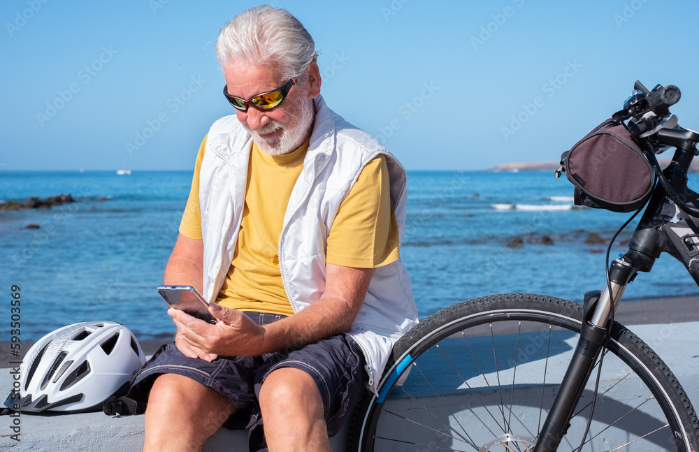 Fototapeta premium Smiling active senior man with electric bicycle on a sunny day sits and talks on the phone. Authentic retirement life and healthy lifestyle concept. Horizon over the sea