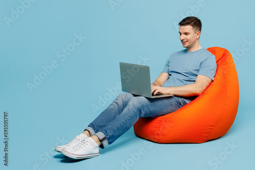 Full body young smiling happy IT man wears casual t-shirt sit in bag chair hold use work on laptop pc computer isolated on plain pastel light blue cyan background studio portrait. Lifestyle concept