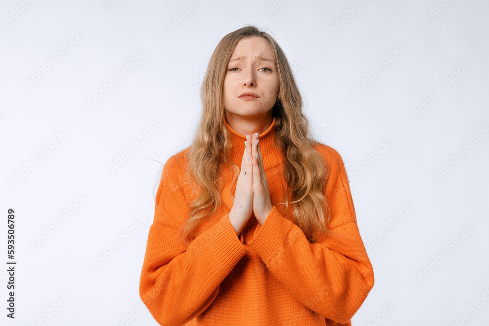 Pleading begging beautiful attractive woman wearing orange sweater standing holding hands folded in prayer looking camera isolated on neutral studio background