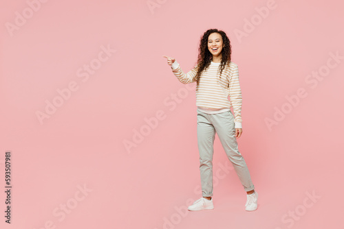 Full body side view happy young woman of African American ethnicity she wear light casual clothes walking going strolling point finger aside isolated on plain pastel pink background studio portrait.