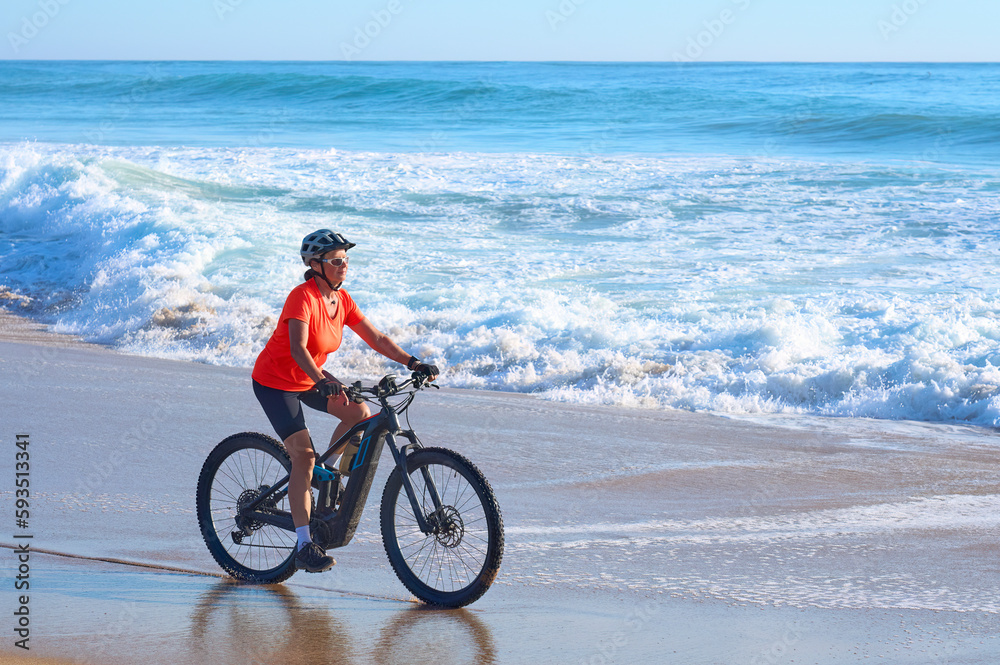 Happy senior woman cycling with her electric mountain bike at the beach of Cape Trafalgar, Costa de la Luz, Andalusia, Spain