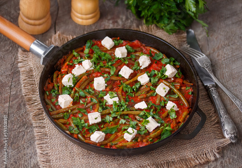 Greek Green beans stew Fasolakia. Tender green beans braised in olive oil and tomato sauce served in cast iron skillet with feta cubes and fresh herbs. Selective focus, horizontal, wooden background. photo