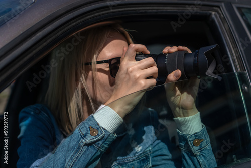 Woman in sunglasses with a camera sits in a car and takes pictures with a professional camera, a private detective or a paparazzi spy. Journalist is looking for sensations and follows celebrities. © Volha