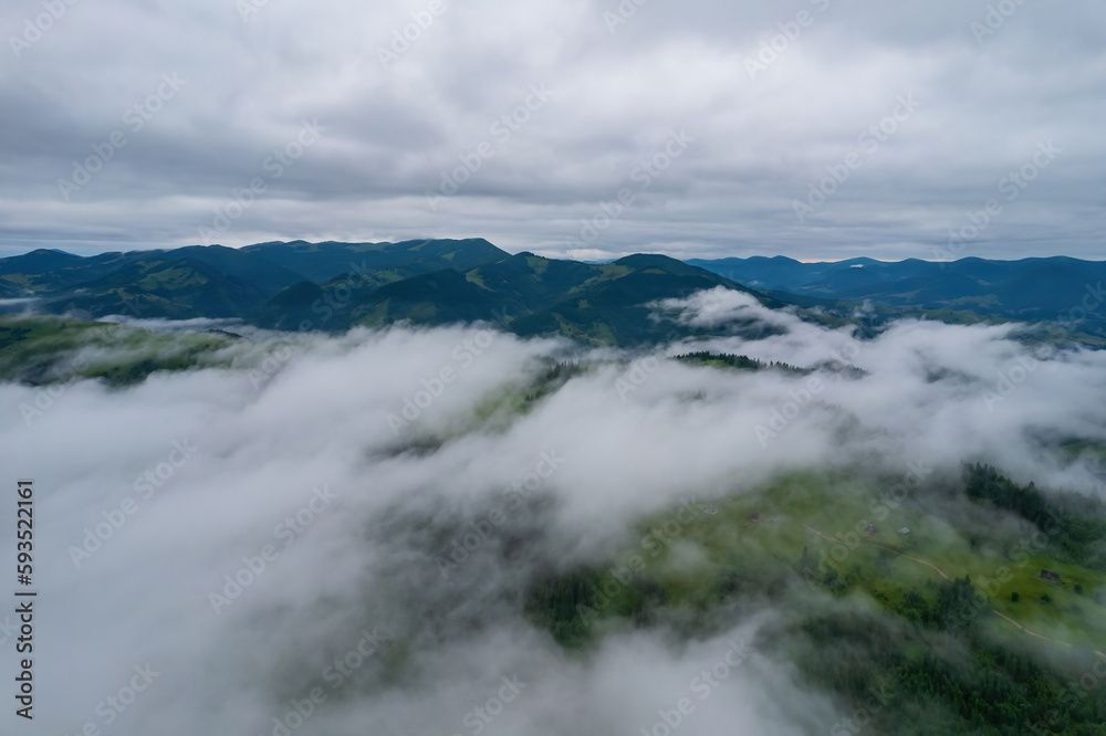 Mountains in clouds at sunrise in summer. Aerial view of mountain peak with green trees in fog. Beautiful landscape with high rocks, forest, sky.