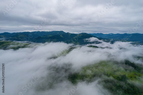 Fototapeta Naklejka Na Ścianę i Meble -  Mountains in clouds at sunrise in summer. Aerial view of mountain peak with green trees in fog. Beautiful landscape with high rocks, forest, sky.